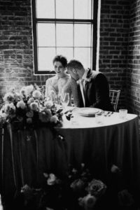 black and white photo of groom kissing brides shoulder at reception table