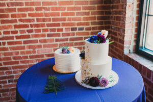 wedding cake with purple and blue flowers sitting on table with blue table cloth